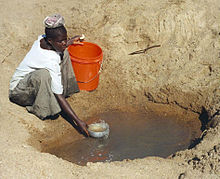 woman getting water out of a hole in the sand and putting it in a bucket.